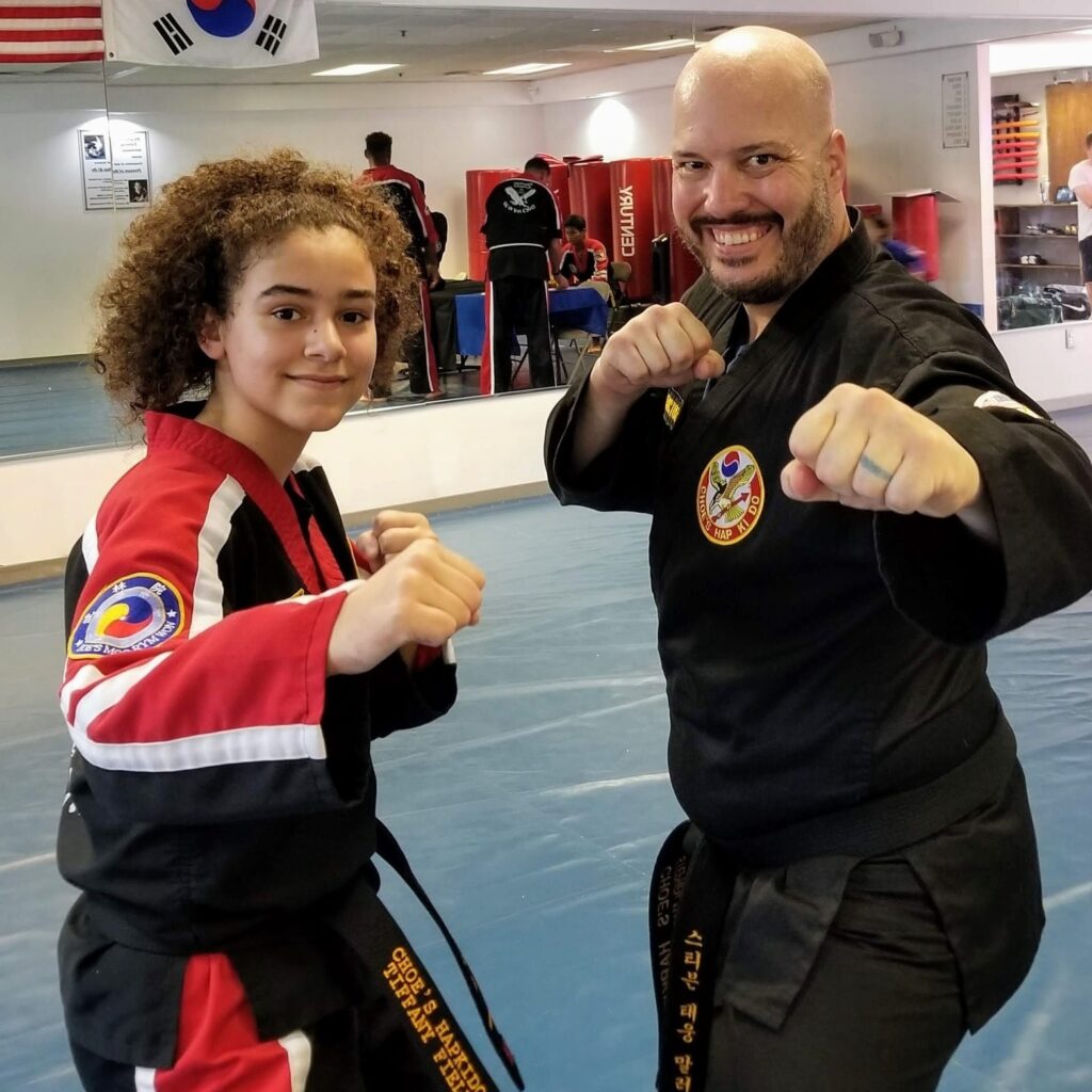 Two martial artists in black and red uniforms pose with fists raised under Korean flag at Choe’s HapKiDo dojo in Cumming, GA.