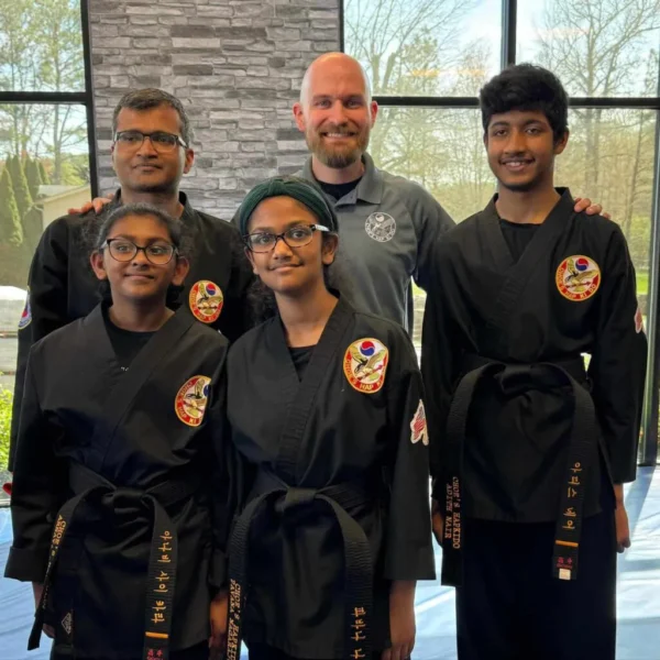 A group of martial arts students and their coach smiling together in front of a brick wall at Choe’s HapKiDo Taneytown, MD.