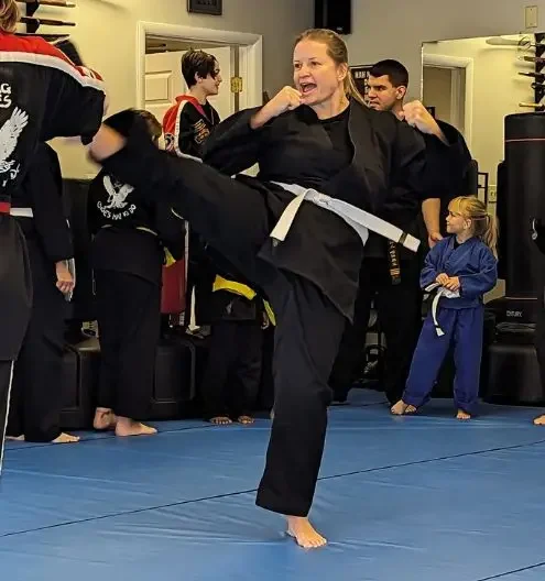 Woman in black martial arts uniform with white belt performing high kick in dojo at Choe’s HapKiDo Flowery, GA, with child observing on blue mats.