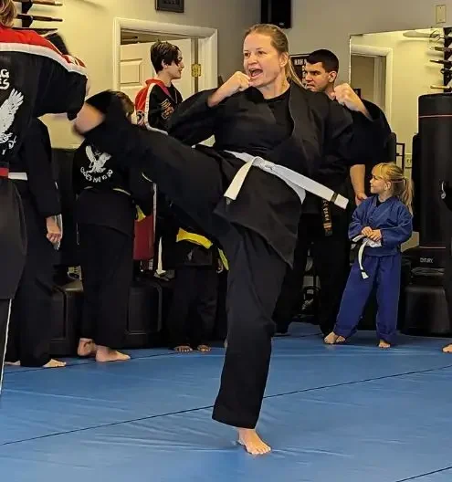 Woman in black martial arts uniform with white belt performing high kick in dojo at Choe’s HapKiDo Duluth, GA, with child observing on blue mats.